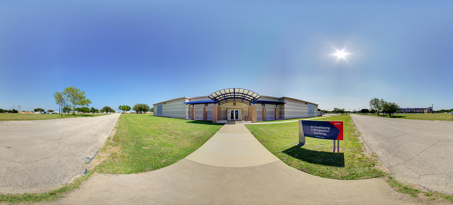 Panoramic view of a single-story campus building with a covered entrance and glass doors, approached by a straight sidewalk, with a nearby sign reading “Air Conditioning & Refrigeration Technology,” surrounded by open grassy areas and parking lots under a bright sunny sky.
