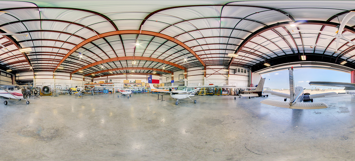 Panoramic view inside an aircraft hangar with multiple small planes parked on a polished concrete floor, maintenance equipment and tools along the walls, and a large open hangar door revealing the runway and bright sky outside.