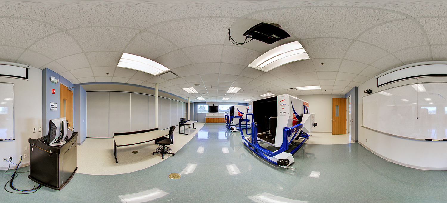 Panoramic view of a classroom with a driving or heavy equipment simulator mounted on a blue platform, facing a projection screen, with desks, chairs, whiteboards, and a ceiling-mounted projector in a clean, well-lit training environment.