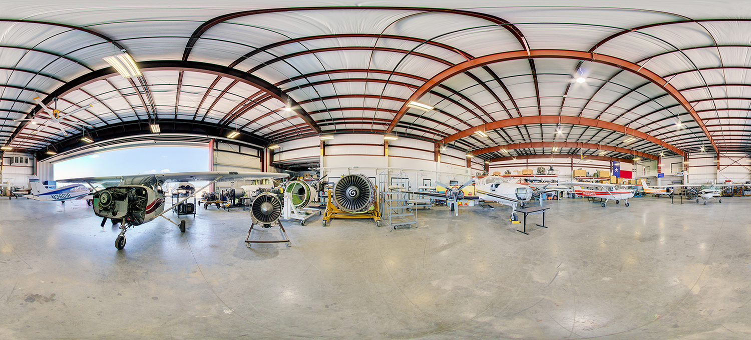 Panoramic view inside an aviation training hangar with several small aircraft, exposed engines and turbine components on stands, maintenance equipment, and a polished concrete floor beneath a high metal-beam ceiling, with an open hangar door letting in daylight.