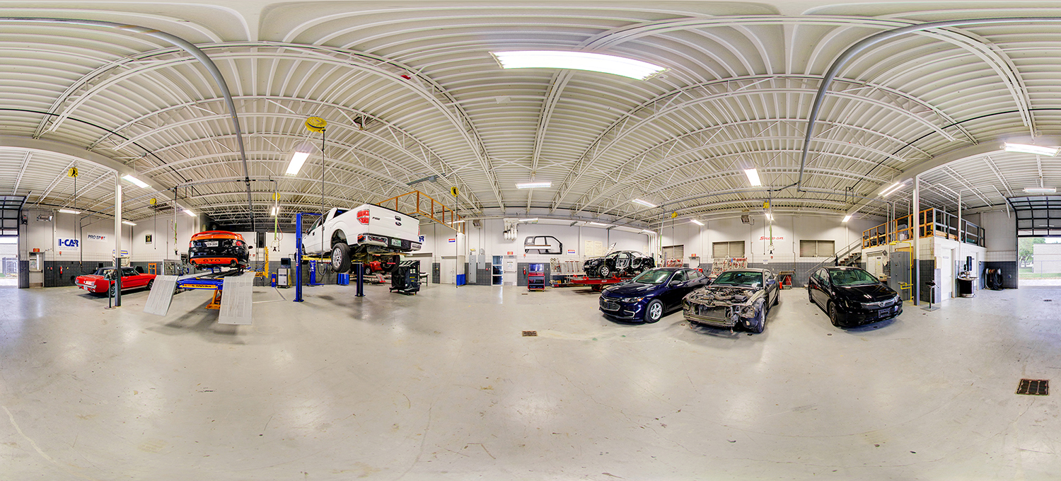 Panoramic view of an automotive training shop with multiple cars and trucks on lifts, including vehicles in various stages of repair, surrounded by workstations, tools, and open floor space under a high, bright ceiling.