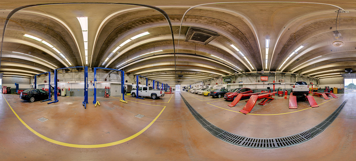 Panoramic view of an automotive training workshop with multiple vehicle lifts, cars and trucks in service bays, red alignment racks, and tool stations spread across a large industrial garage with a high concrete ceiling and bright overhead lighting.