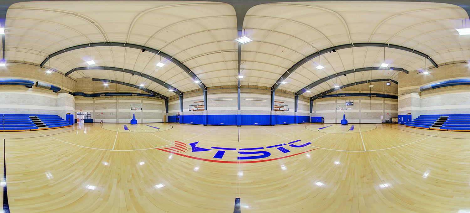 Panoramic view of an indoor basketball gymnasium with a polished wood court featuring an “TSTC” logo at center, multiple hoops, blue wall padding, and bleachers along the sides under a high arched ceiling.