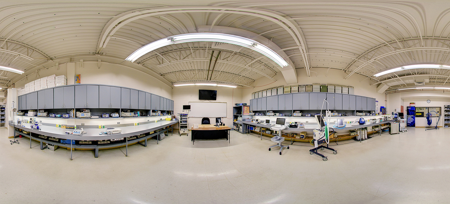 Panoramic view of a technical lab classroom with long workbenches lined with electronic testing equipment and medical devices, overhead storage cabinets, a central instructor desk, and bright industrial lighting in a clean, organized training space.