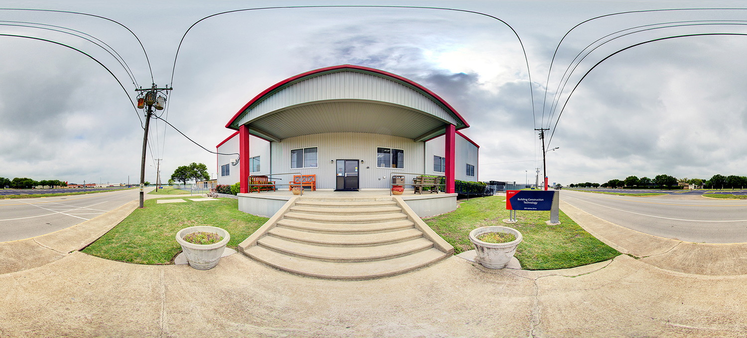 Panoramic view of a small campus building labeled “Building Construction Technology,” featuring a raised entrance with steps, red support columns, and a curved roof, set beside a road with grassy areas, utility poles, and a cloudy sky overhead.