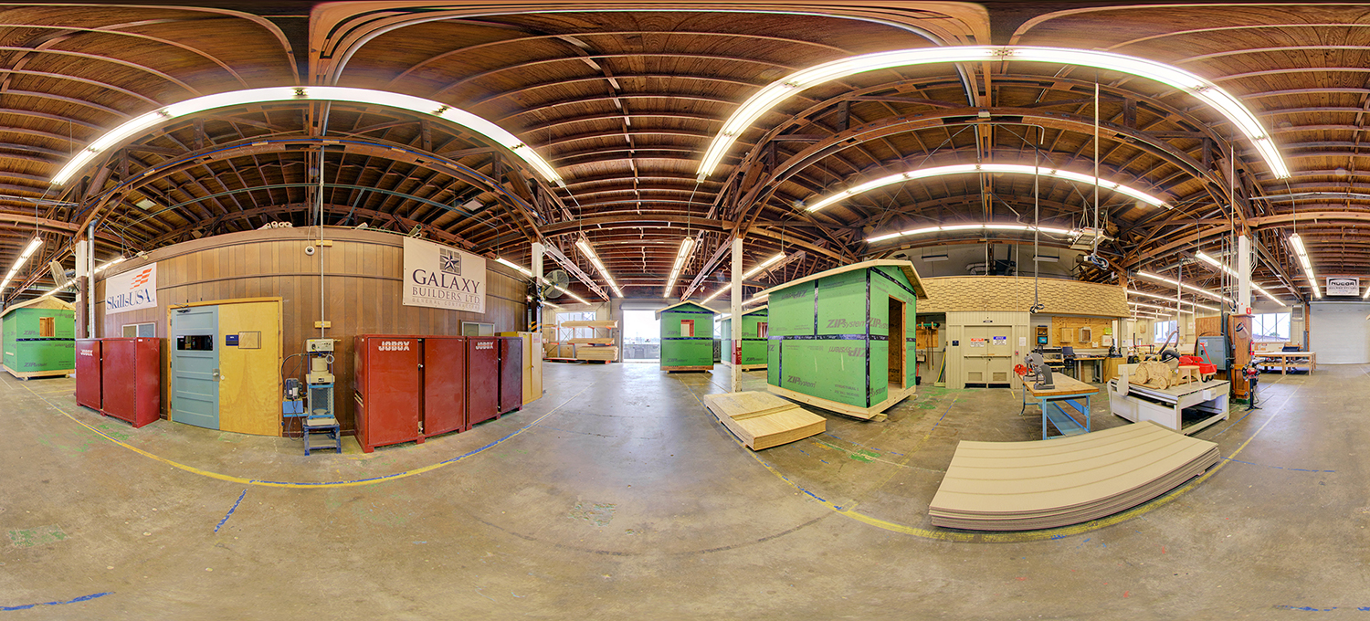 Panoramic view of a construction training workshop with wood-framed practice structures, tool cabinets, and workstations, set under a high ceiling with exposed beams and bright industrial lighting.