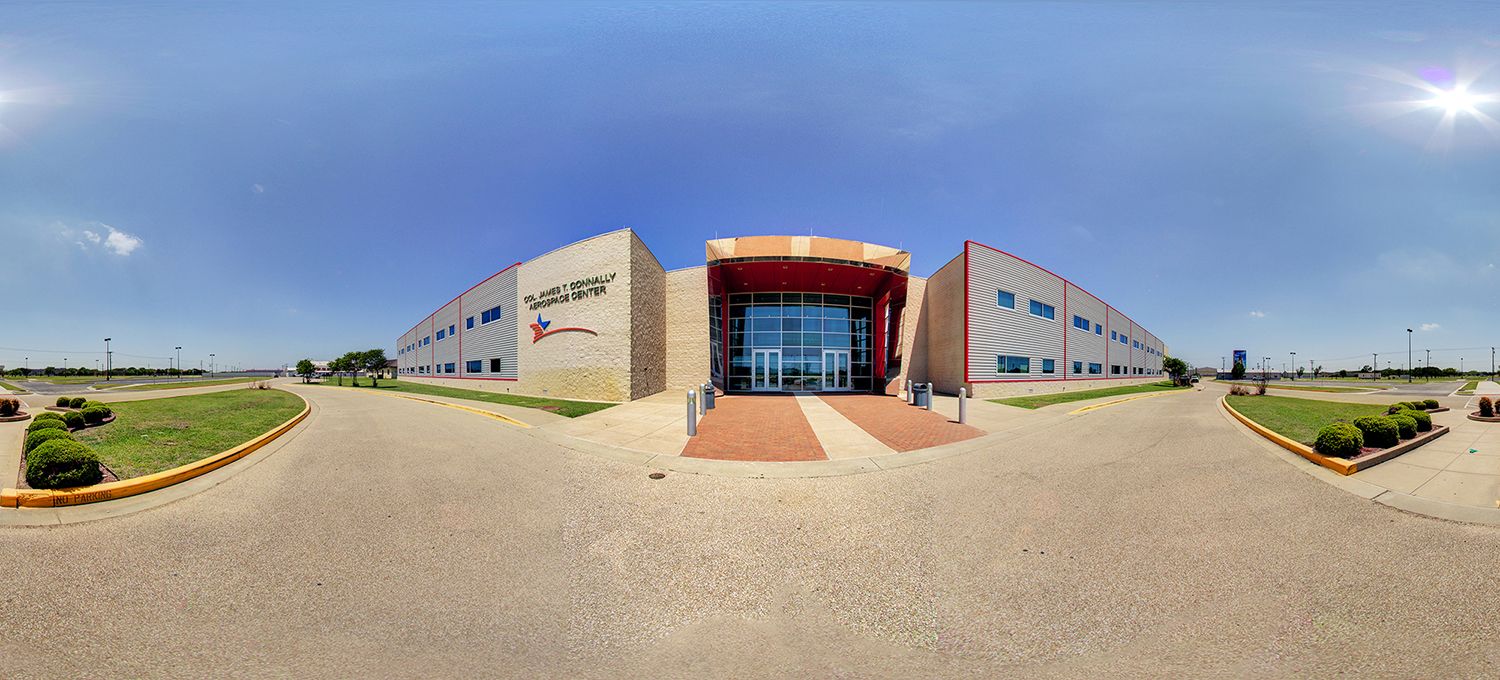 Panoramic view of the Dr. Col James T Connally Aerospace Center, featuring a modern symmetrical building with a glass-front entrance, red architectural accents, and surrounding driveways and landscaped areas under a clear, sunny sky.