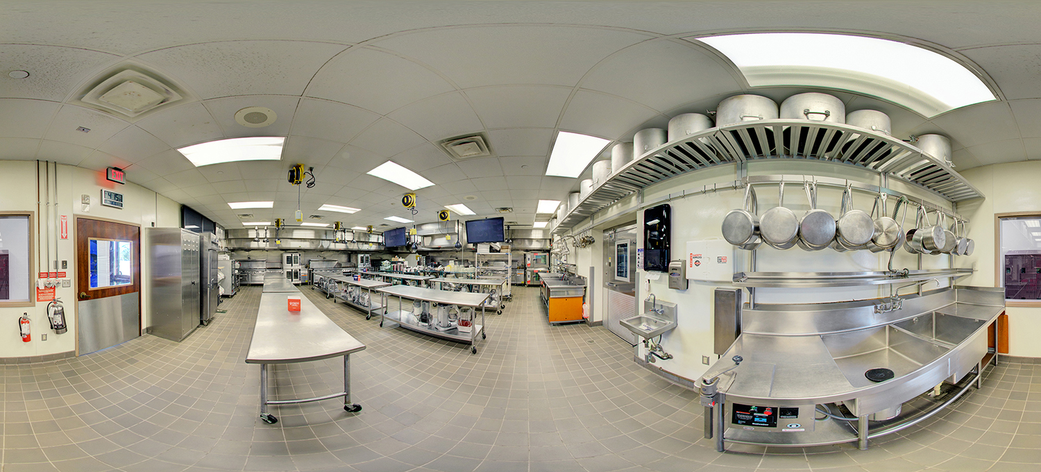 Panoramic view of a commercial teaching kitchen with stainless steel prep tables, hanging pots and pans, large sinks, industrial ovens, and cooking stations arranged across a tiled floor under bright ceiling lights.