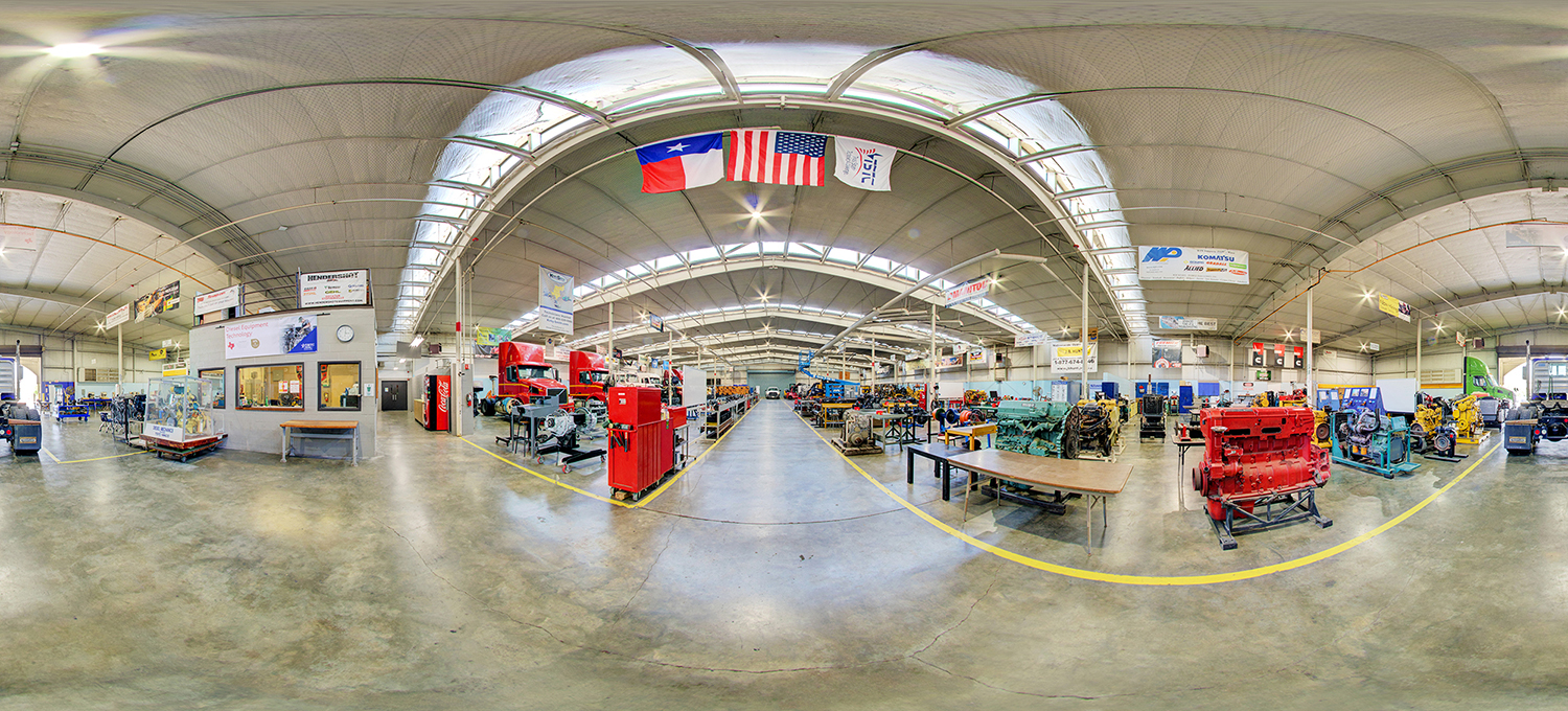Panoramic view of a large industrial training workshop filled with engines, heavy equipment components, and workstations arranged in rows, with tool cabinets, trucks, and machinery under a high arched ceiling, and U.S. and Texas flags hanging overhead.