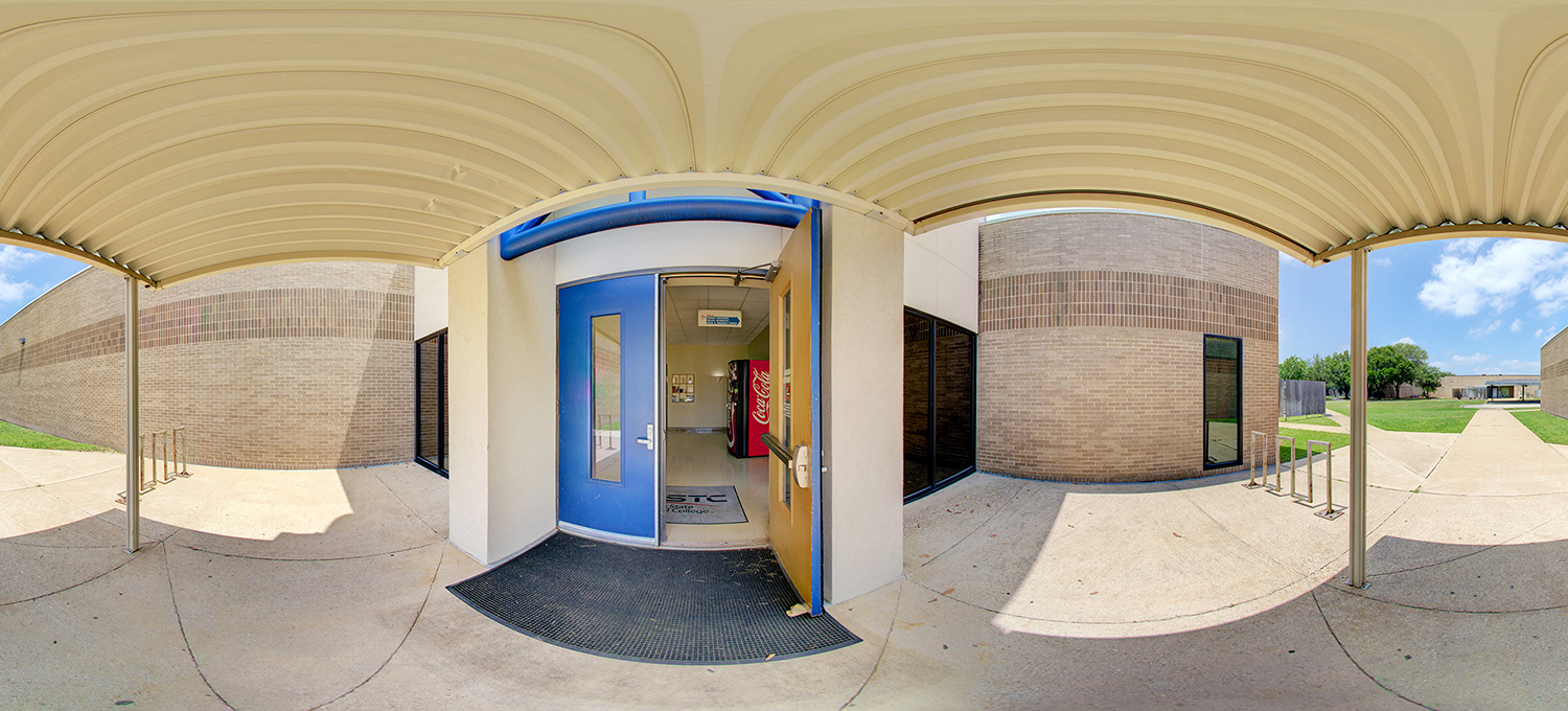 Panoramic view of a covered outdoor walkway leading to a building entrance with a blue door propped open, showing a glimpse of an interior hallway with a vending machine, surrounded by brick walls, bike racks, and a sunny campus courtyard.