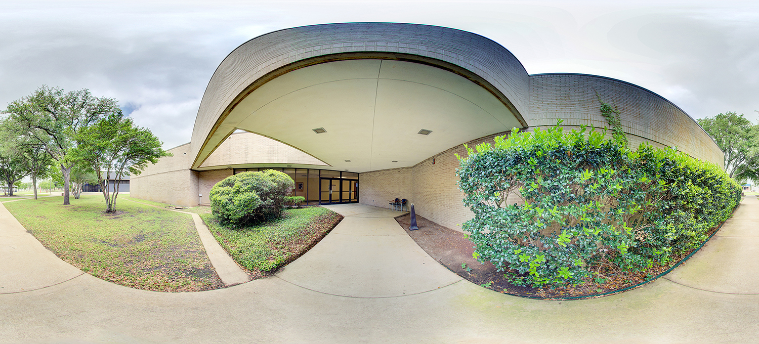 Panoramic view of a building entrance with a curved overhang, glass double doors, and surrounding landscaped shrubs and trees, set along a concrete walkway under an overcast sky.