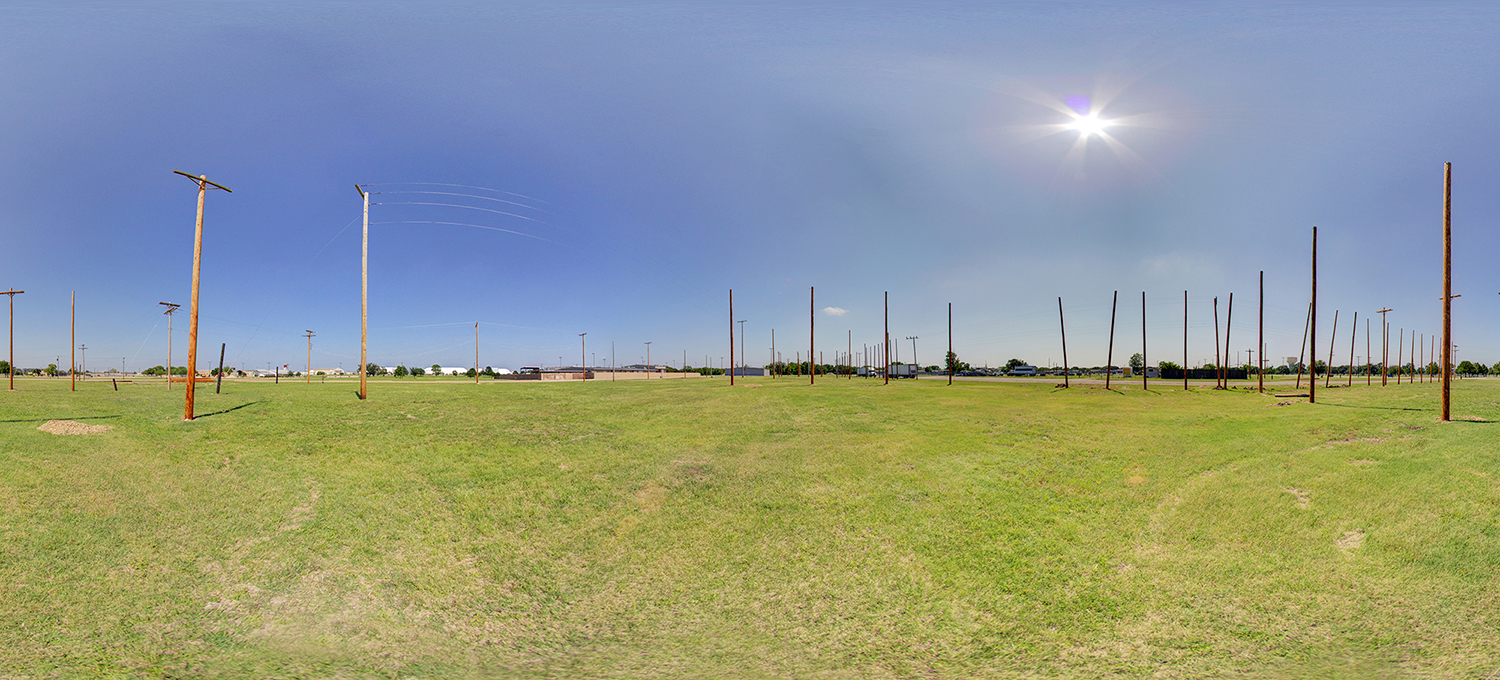 Panoramic view of an open grassy training field with numerous utility poles arranged in rows for electrical linework practice, under a clear blue sky with bright sunlight and minimal surrounding buildings.