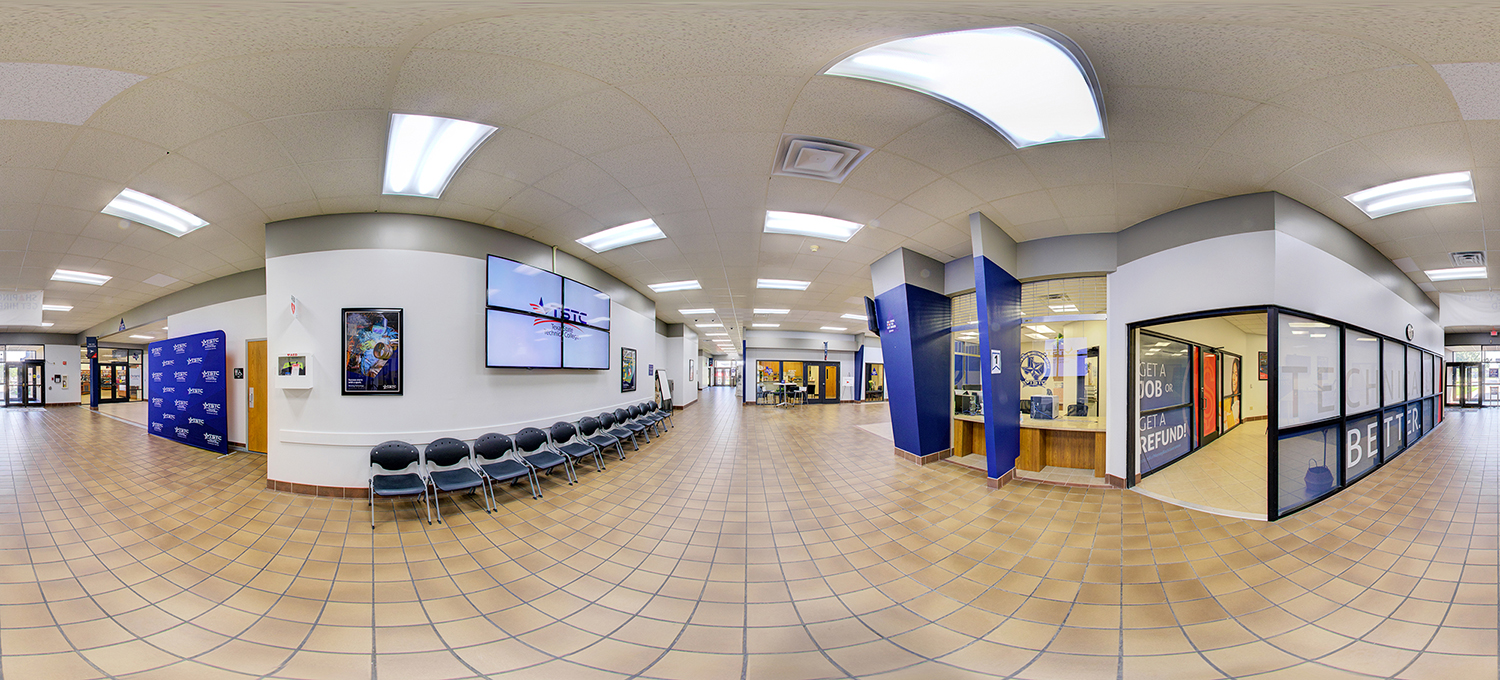 Panoramic view of a campus lobby featuring tiled floors, a row of waiting chairs beneath large wall-mounted screens, a glass-front service office, and hallways leading to classrooms and offices.
