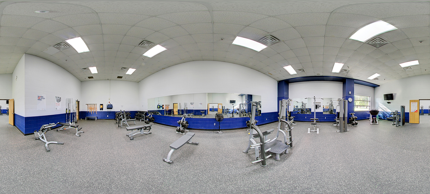 Panoramic view of a fitness room with weight machines, benches, free weights, and exercise equipment arranged across a carpeted floor, with mirrored walls and bright overhead lighting.