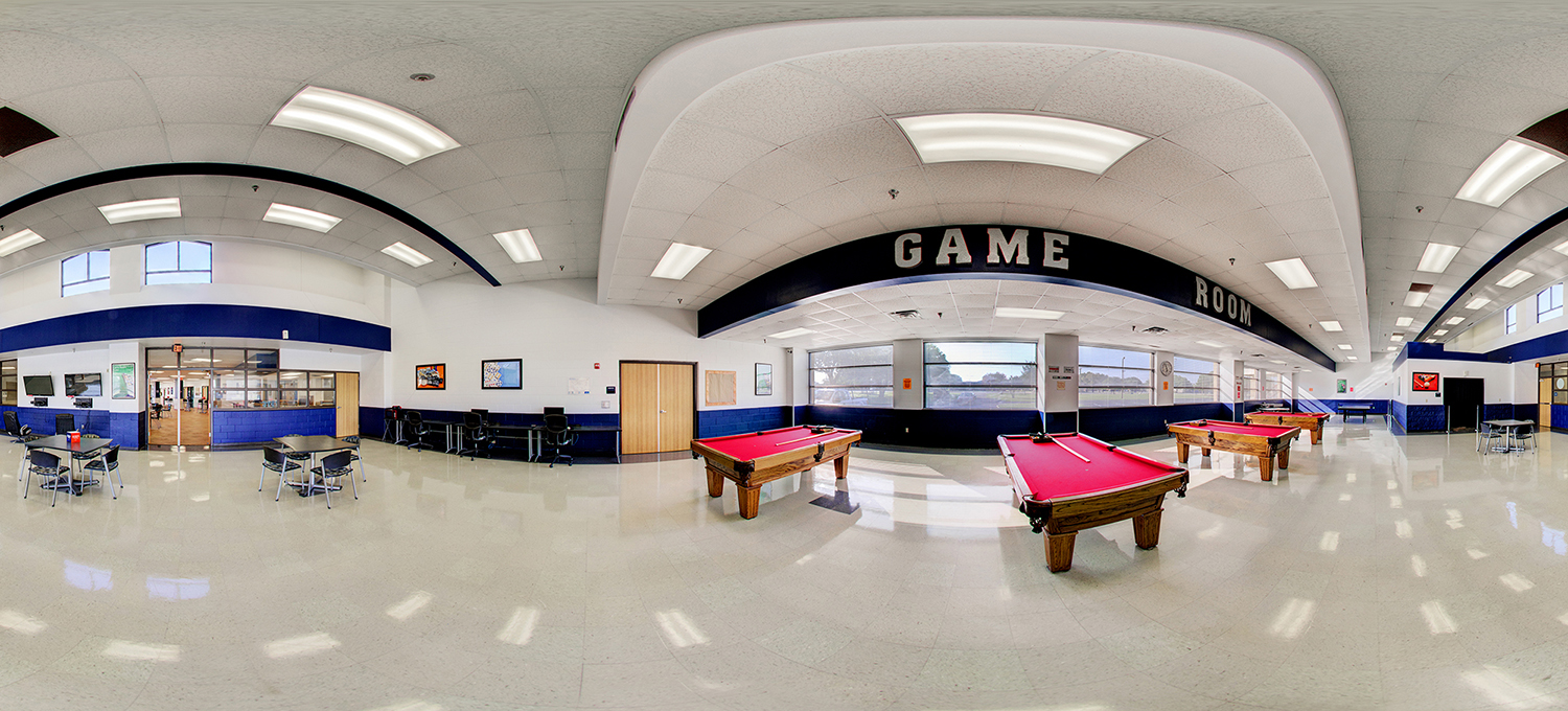 Panoramic view of a campus game room featuring multiple red-felt pool tables, seating areas with tables and chairs, computers along one wall, and large windows bringing in natural light beneath a “GAME ROOM” sign.