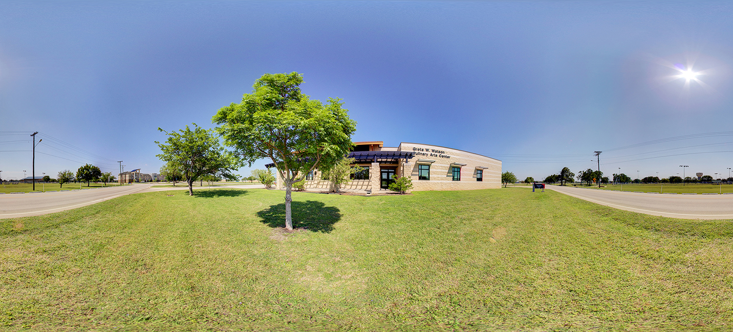 Panoramic view of a small campus building labeled “Culinary Arts Center,” set on a grassy lawn with a leafy tree in the foreground, clear blue sky overhead, and surrounding roads on either side.