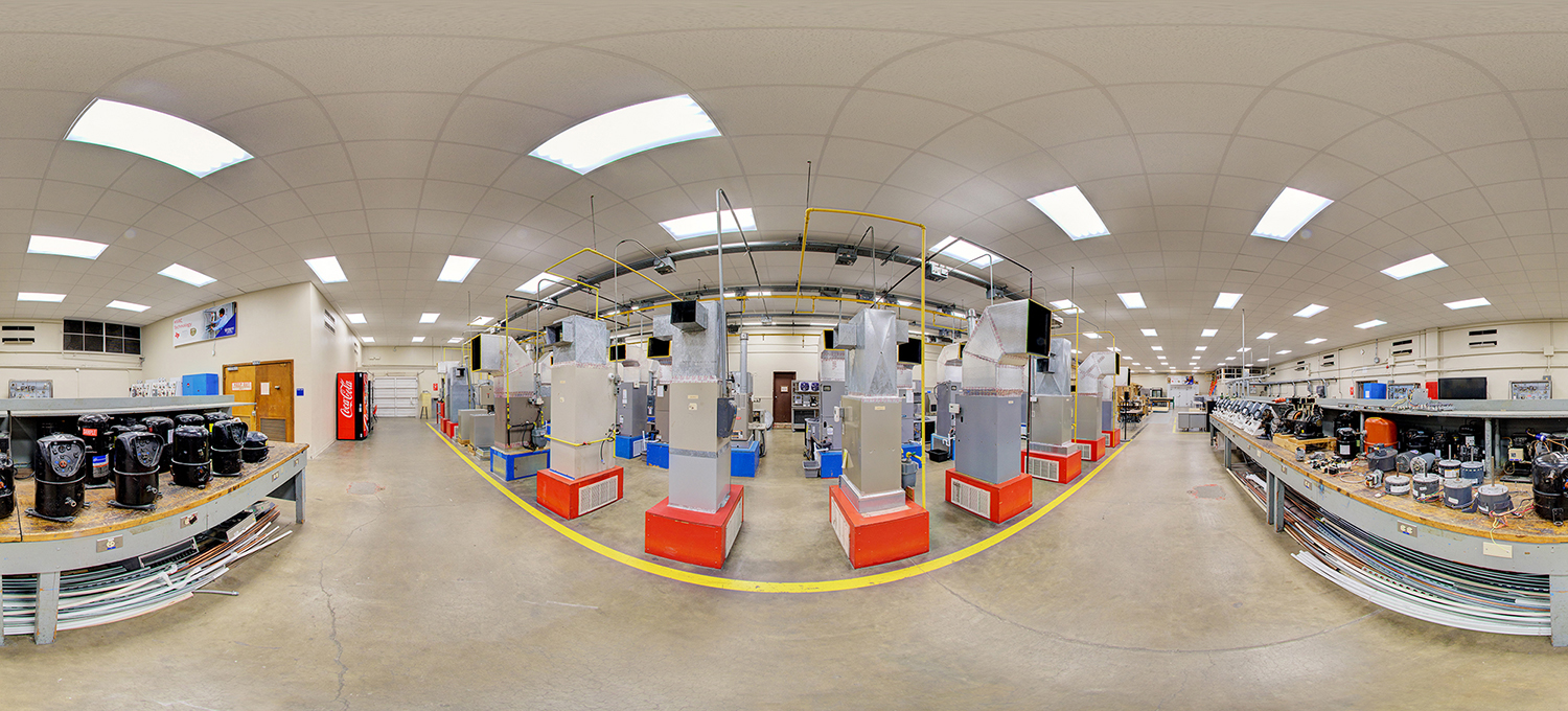 Panoramic view of an HVAC training lab filled with rows of large ventilation units and duct systems, surrounded by workbenches with compressors and tools, all under bright fluorescent lighting in a spacious classroom setting.