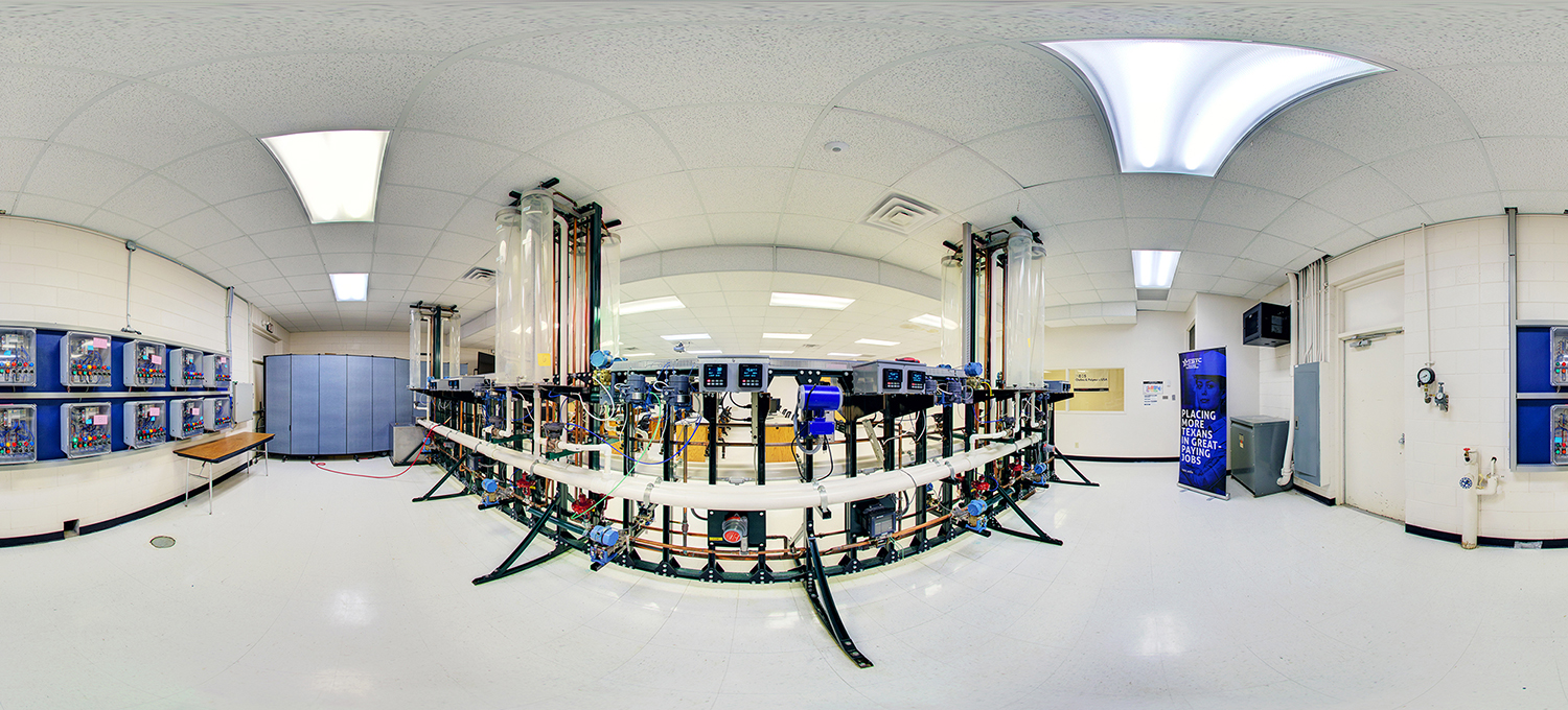 Panoramic view of an industrial process control lab featuring a central circular system of pipes, valves, gauges, and digital control panels, surrounded by instructional stations and equipment, in a bright, clean technical training environment.
