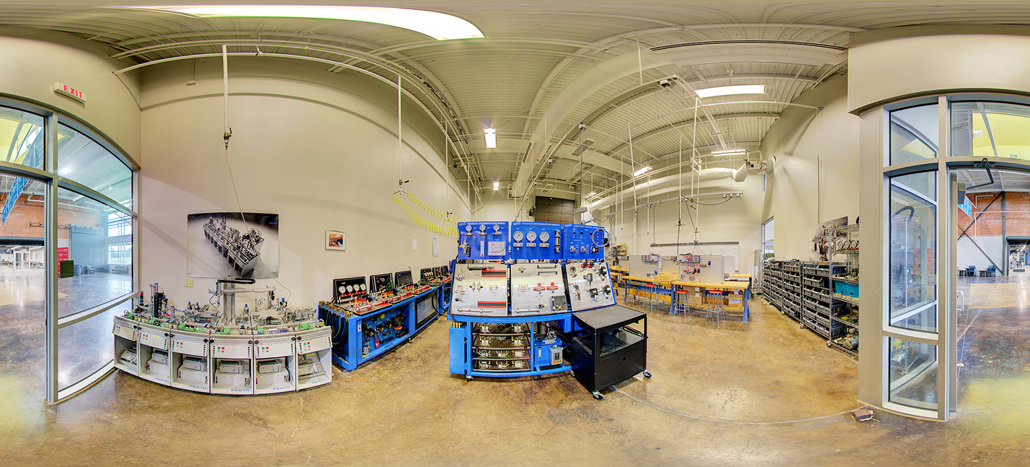 Panoramic view of a technical training lab with blue pneumatic and hydraulic training stations, gauges and control panels, workbenches, and organized parts shelves inside a high-ceiling industrial classroom with exposed piping and bright lighting.