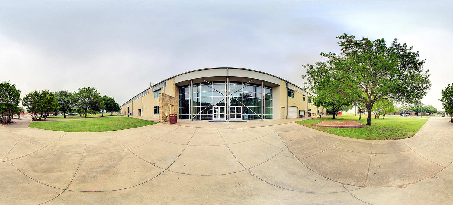 Panoramic view of a modern campus building with a curved glass entrance façade, surrounded by sidewalks, green lawns, and trees under an overcast sky.