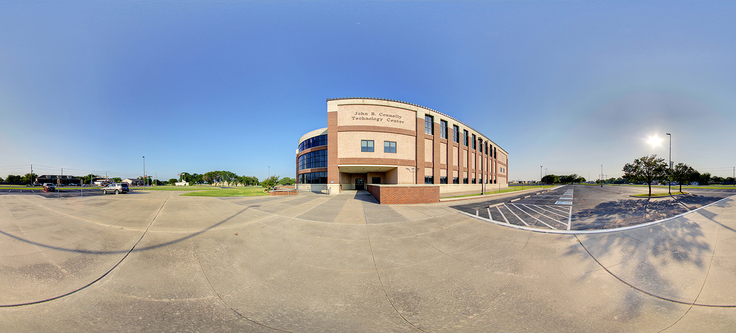 Panoramic view of a large academic building labeled “John B. Connally Technology Center,” featuring a curved glass corner, brick façade, and surrounding parking areas under a clear sunny sky.