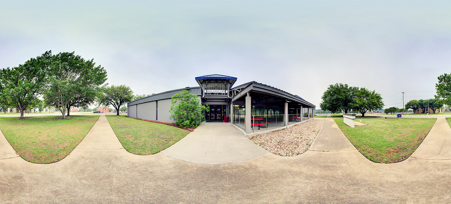 Panoramic view of a low, modern campus building with a covered outdoor seating area, glass entrance, and surrounding sidewalks and trees on a grassy lawn under an overcast sky.