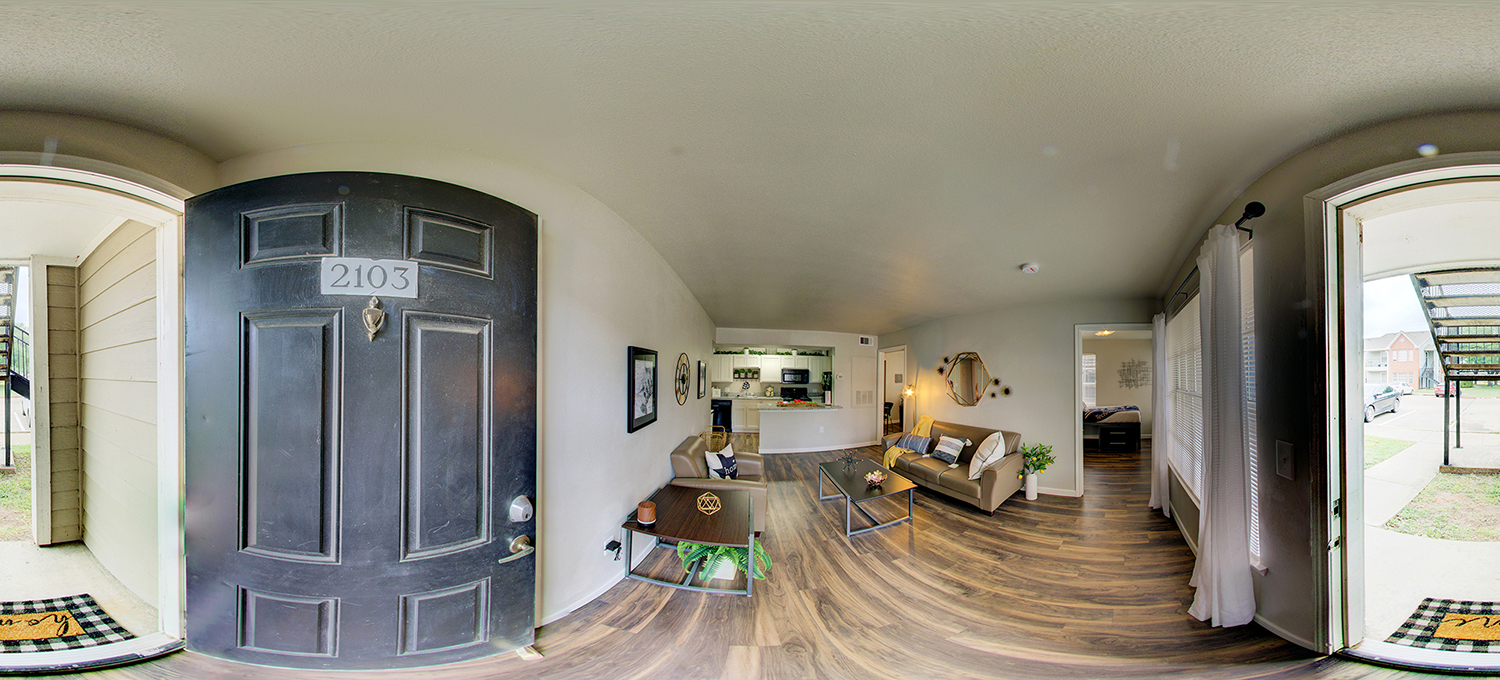 Panoramic view from an apartment entryway showing a black door labeled “2103,” opening into a modern living room with wood flooring, a sofa and coffee table, a small dining area, and a kitchen in the background, with a bedroom visible down a hallway.