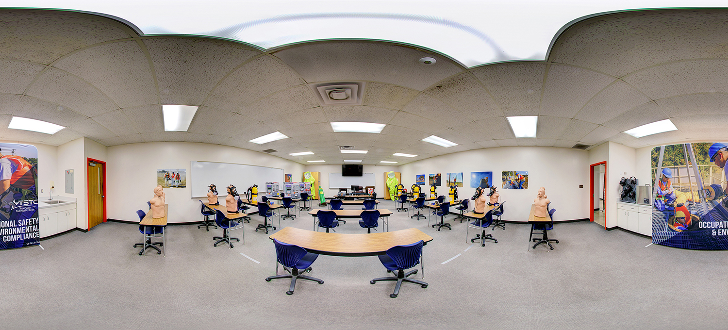 Panoramic view of a safety training classroom with curved rows of desks and chairs, CPR mannequins placed at each station, and protective gear and equipment displayed along the walls beneath instructional posters.