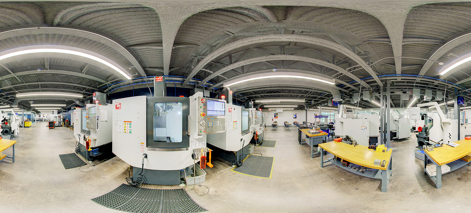Panoramic view of an advanced manufacturing lab with multiple CNC machines, control panels, and worktables arranged across a spacious industrial room with exposed ceiling beams and bright overhead lighting.