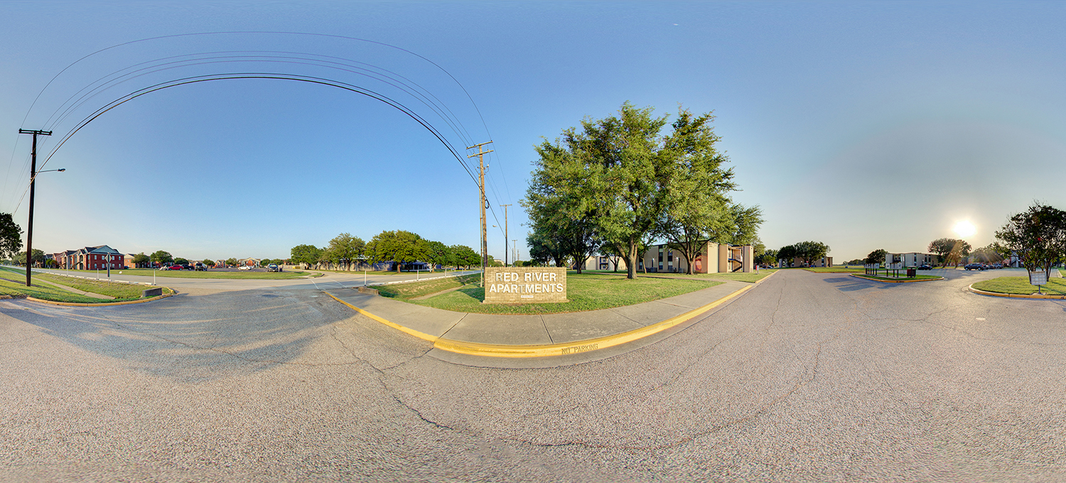 Panoramic view of an apartment complex entrance with a “Red River Apartments” sign on a grassy median, surrounded by low-rise residential buildings, sidewalks, and a roadway under a clear sky with the sun low on the horizon.