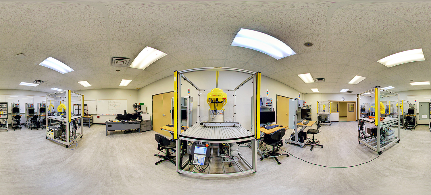 Panoramic view of a robotics lab with multiple industrial robotic arms enclosed in safety frames, workstations with computers and control panels, and a central automated system, all set in a bright classroom environment with tiled floors and overhead lighting.