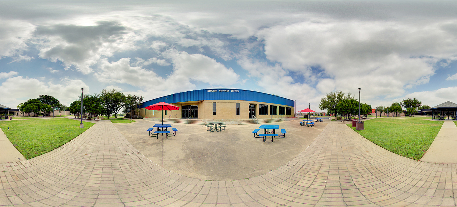 Panoramic view of a campus Student Services Center building with a blue facade, surrounded by paved walkways, grassy areas, and outdoor seating with picnic tables and red umbrellas under a partly cloudy sky.