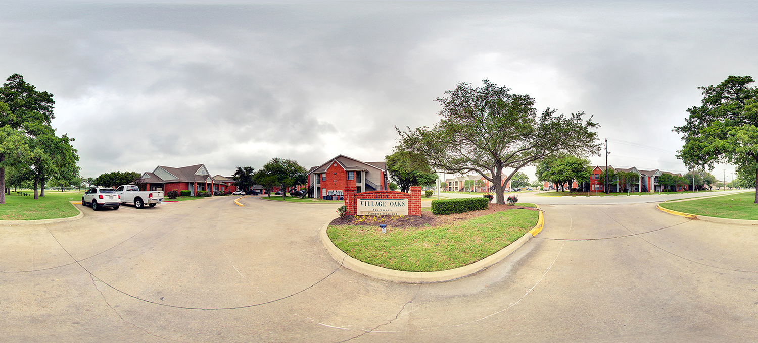 Panoramic view of an apartment complex entrance with a “Village Oaks Apartments” sign on a landscaped median, surrounded by red-brick residential buildings, parked cars, and tree-lined streets under an overcast sky.