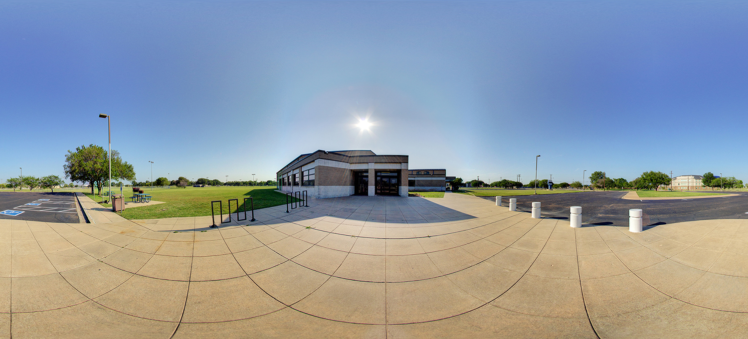 Panoramic exterior view of a campus building entrance with a paved walkway, bike racks, and bollards in front, surrounded by a large parking lot, grassy areas with trees, and clear blue sky with bright sunlight overhead.