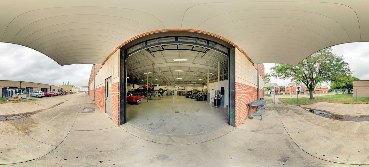 Panoramic view of an open automotive workshop bay with a large garage door, several cars parked inside, vehicle lifts and tools visible, and outdoor parking areas and buildings on either side of the entrance.