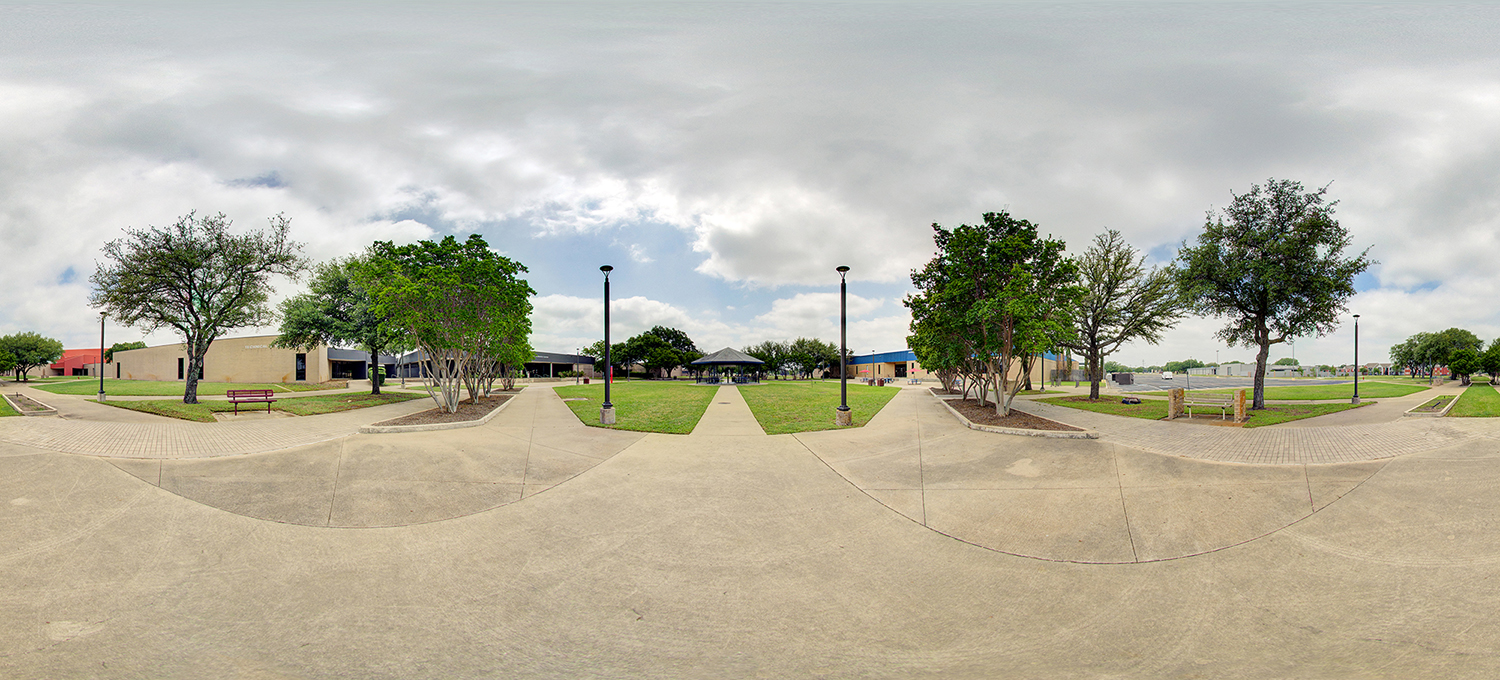 Panoramic view of a landscaped campus courtyard with intersecting concrete walkways, grassy lawns, benches, and evenly spaced trees, leading toward a central gazebo under a cloudy sky.