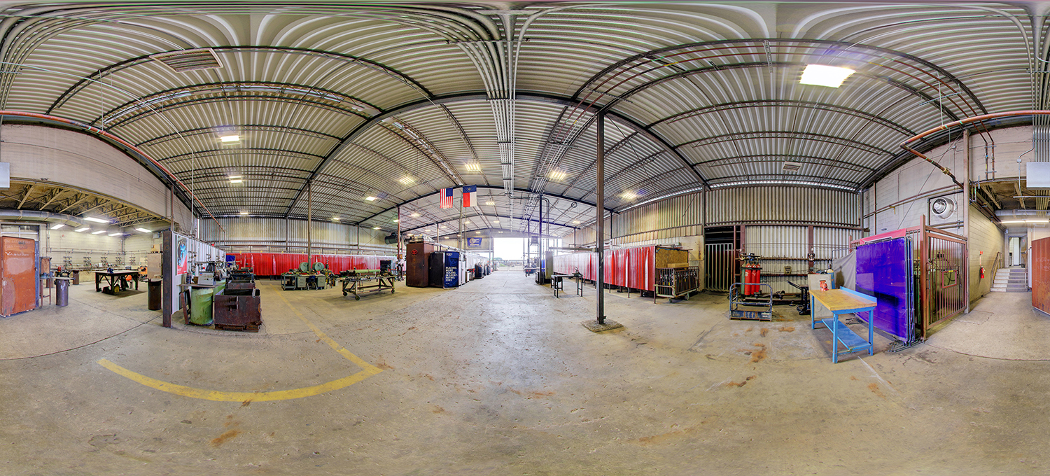 Panoramic view of a large industrial welding shop with rows of red welding booths, metalwork stations, gas cylinders, and worktables spread across a spacious warehouse with a high, ribbed metal ceiling and overhead lighting.