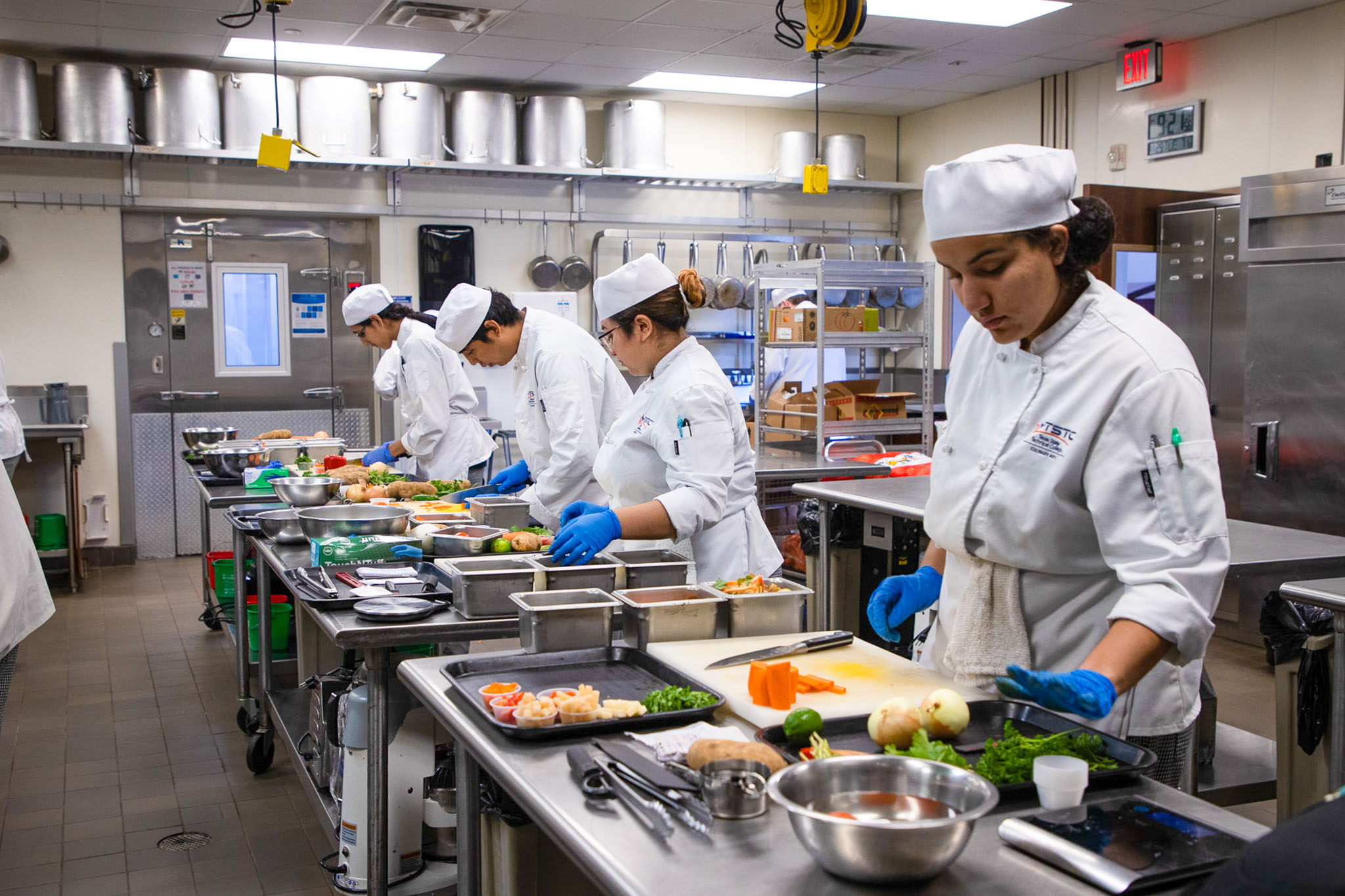 Culinary students in white chef uniforms and blue gloves prepare ingredients at stainless steel workstations, with one student slicing vegetables in the foreground while others chop and assemble food in a commercial kitchen.