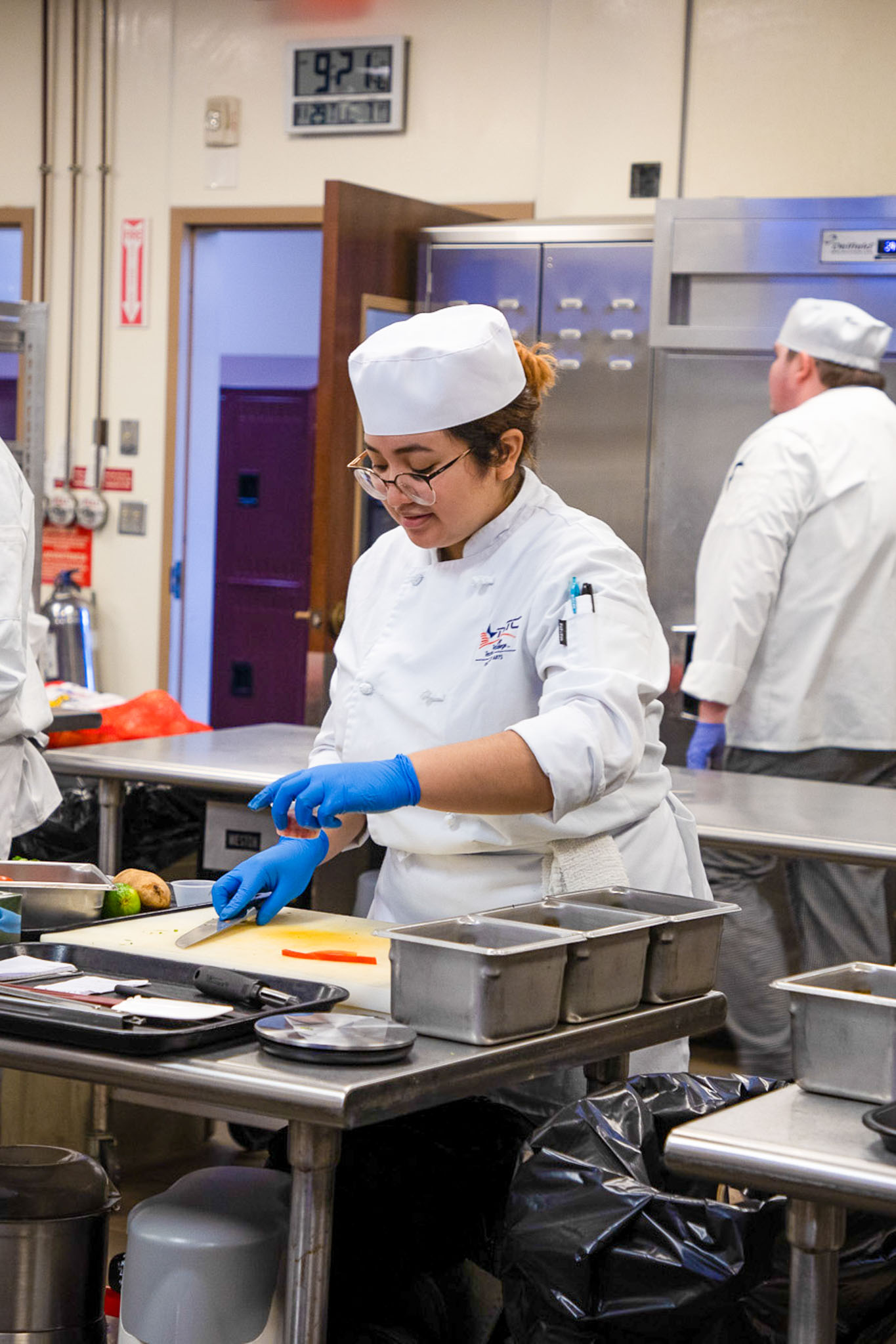 Culinary student in a white chef coat and hat, wearing blue gloves, slices a carrot on a cutting board at a stainless steel workstation in a commercial kitchen, with other chefs working in the background.