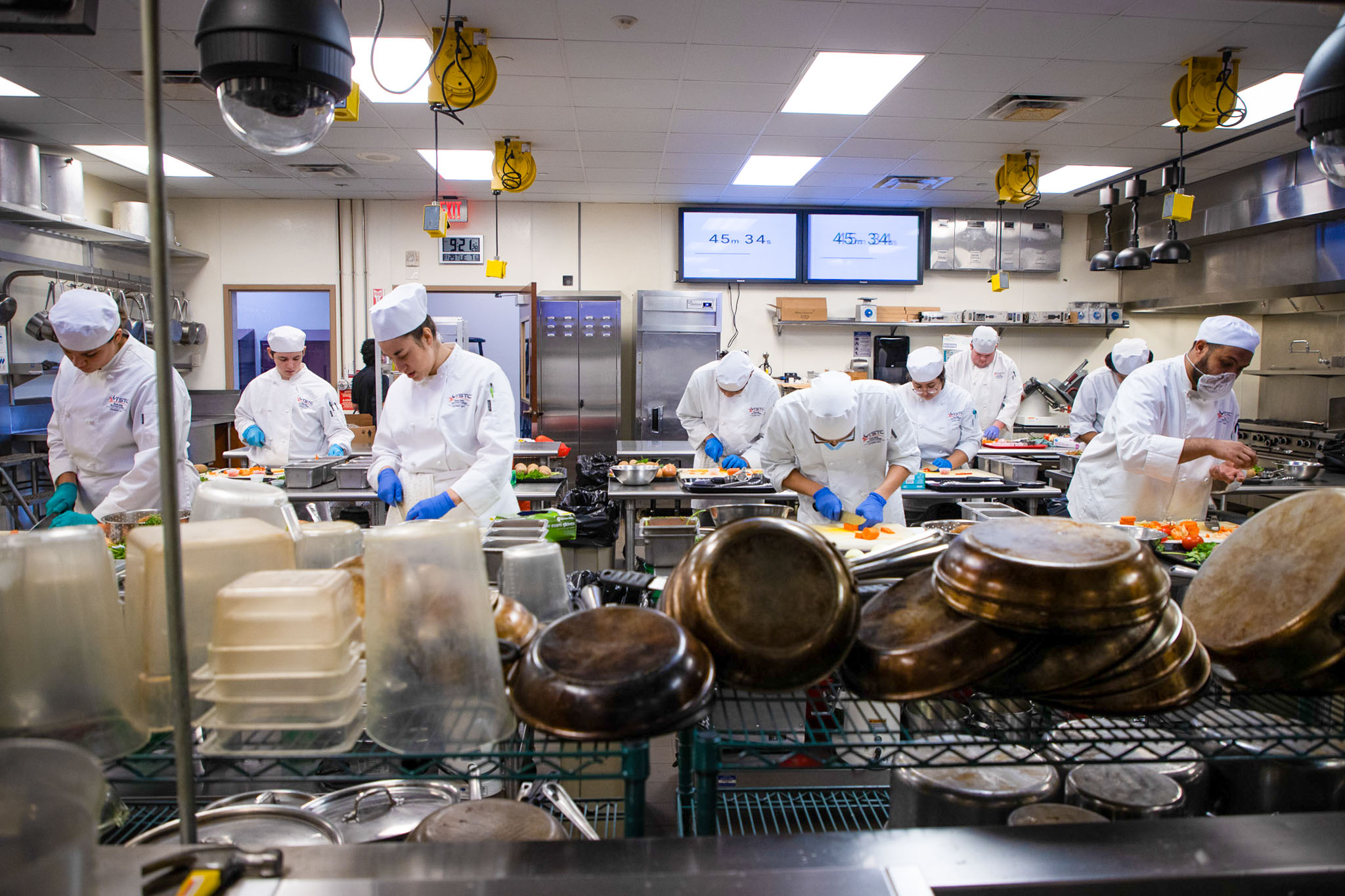 Wide view of a commercial kitchen classroom where multiple culinary students in white chef uniforms and blue gloves work at stainless steel stations, chopping vegetables while stacks of pots and containers sit in the foreground.