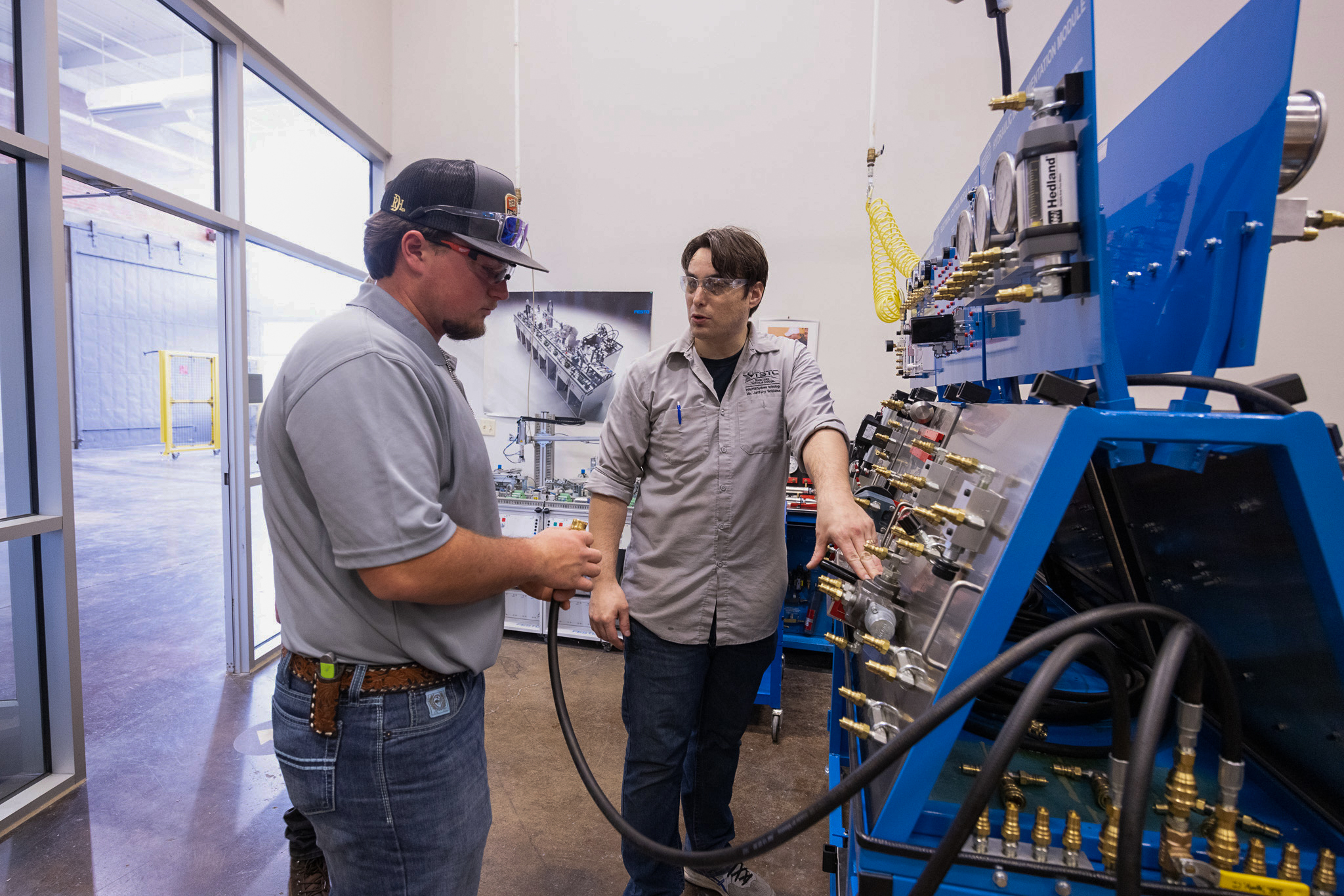 Two students wearing safety glasses work at a hydraulic training panel; one holds a hose fitting while the other points to valves and gauges, explaining the system.