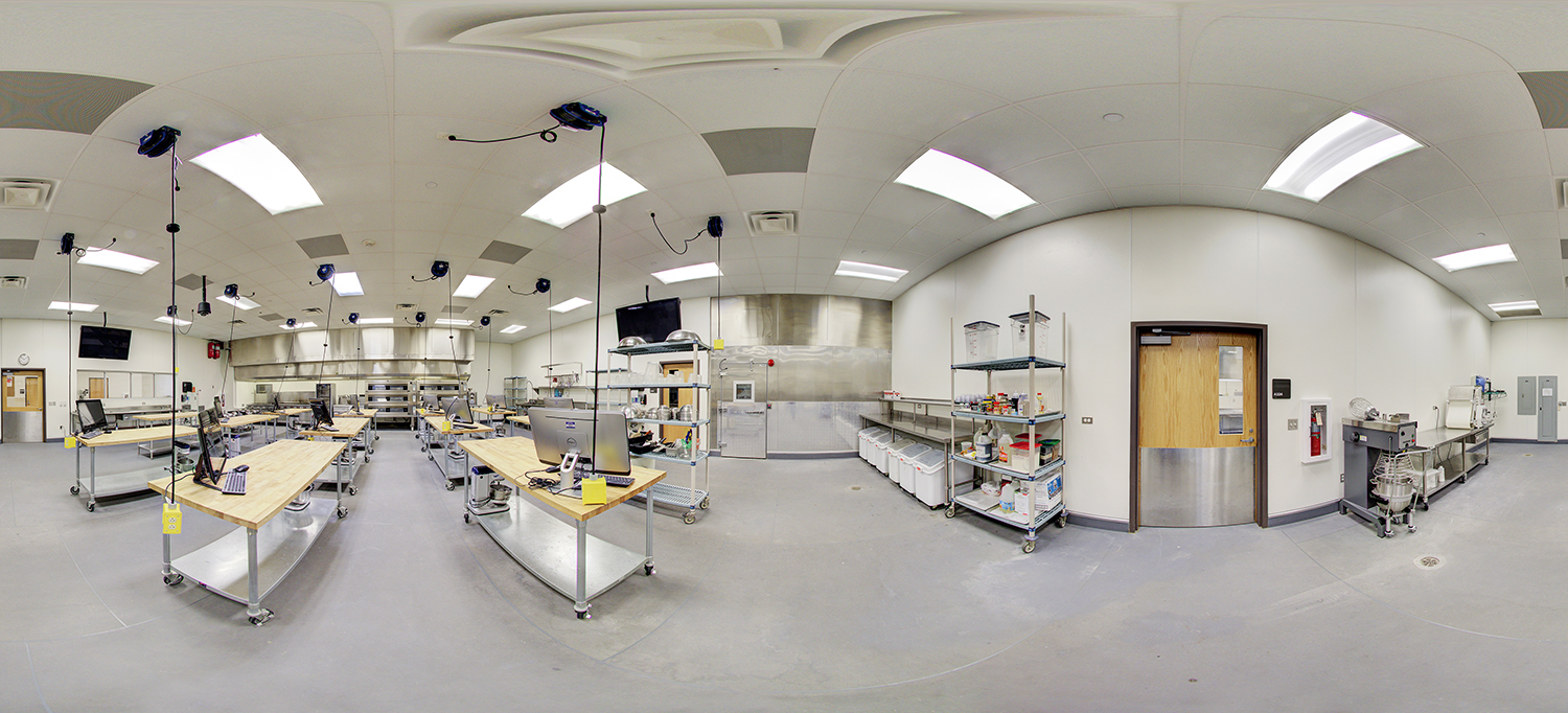 Panoramic view of a culinary training kitchen with wooden prep tables, computer stations, hanging power outlets, stainless steel cooking equipment, storage racks with ingredients, and commercial mixers arranged throughout a clean, well-lit workspace.