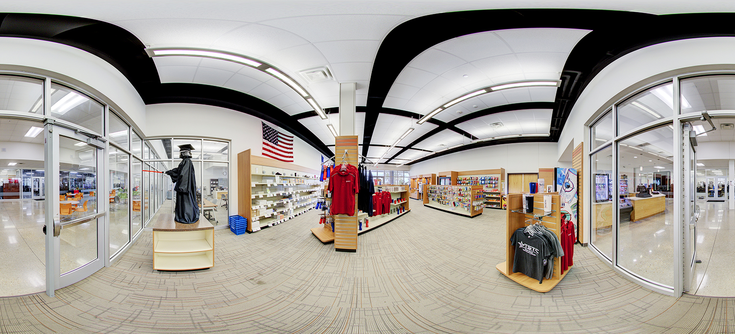 Panoramic view of a campus retail area with shelves of merchandise, including red and gray apparel, accessories, and supplies; a mannequin in graduation attire stands near the entrance, and an American flag hangs on the wall above.