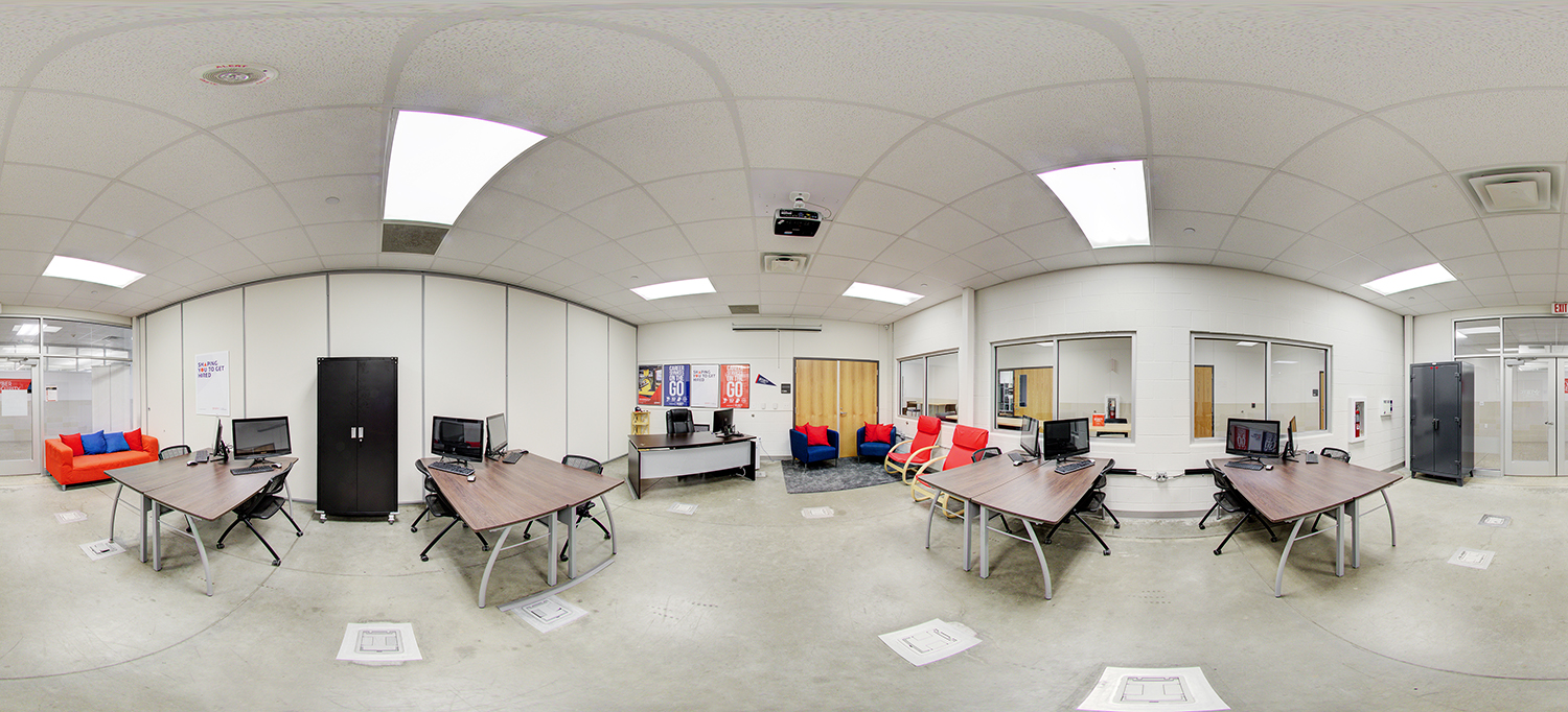 Panoramic view of a modern area with multiple desks equipped with desktop computers and office chairs, a black storage cabinet, and a seating area with red and blue chairs near the back wall, along with posters, a desk, and interior windows facing adjacent rooms.