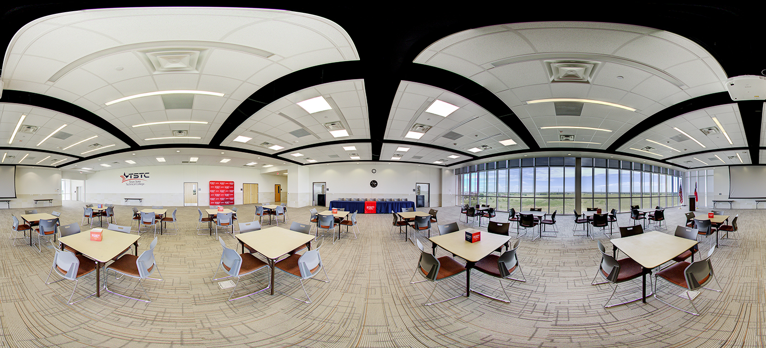 Panoramic view of a large, modern multipurpose room with round tables and chairs arranged across a carpeted floor, a presentation area with tables and a backdrop at the front, and a wall of tall windows letting in natural light with flags displayed nearby.
