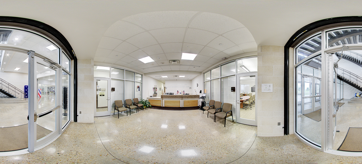 Panoramic view of a reception area with a central front desk, chairs lined along the walls for waiting, glass-walled offices on both sides, and entry doors leading to hallways and adjacent spaces.