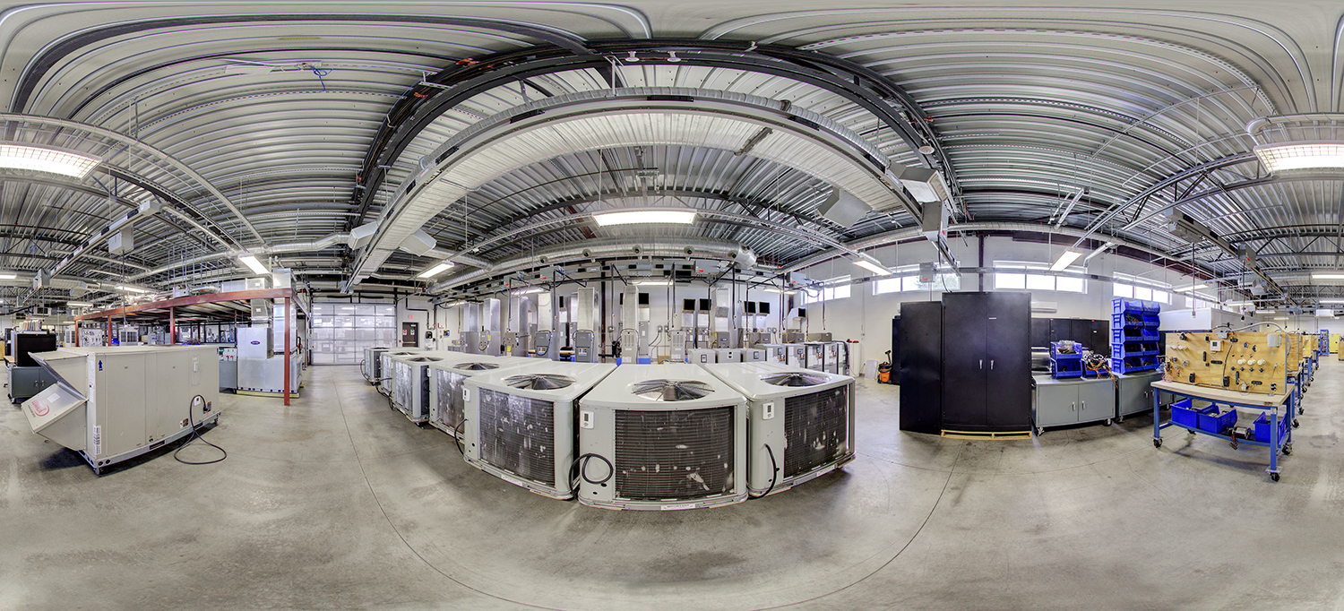 Panoramic view of an industrial training lab with multiple HVAC units arranged in rows, exposed ceiling ductwork, electrical panels, and workstations with tools and components for hands-on technical instruction.