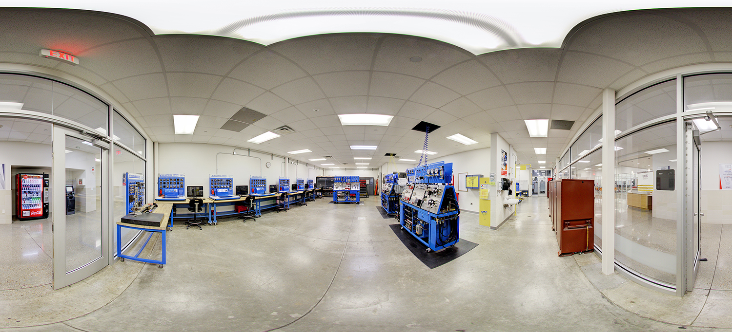 Panoramic view of a technical training lab with blue workstations equipped with mechanical and electrical training panels, tools, and computers, surrounded by glass-walled classrooms and adjacent hallways.