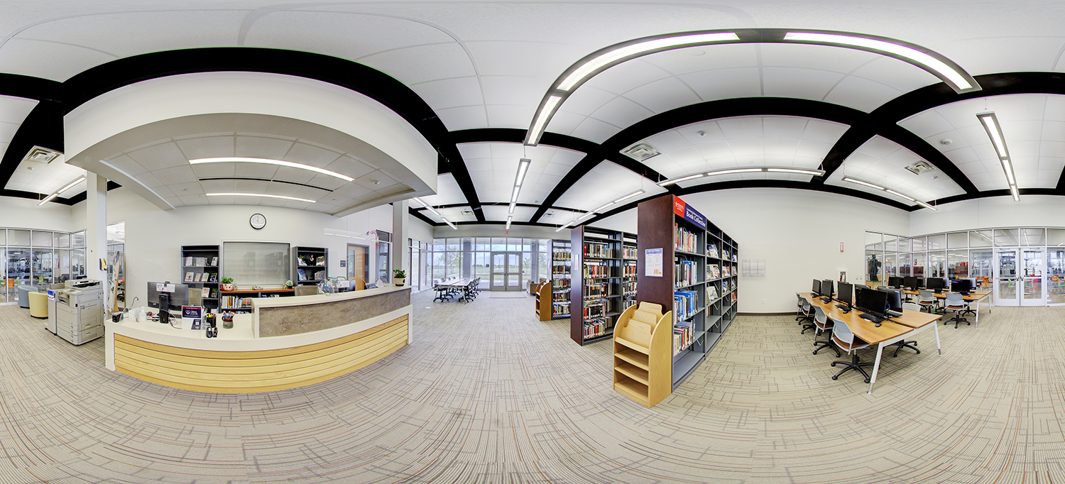 Panoramic view of a library area with a front service desk, bookshelves filled with books, computer workstations along the wall, and study tables near large windows at the far end.