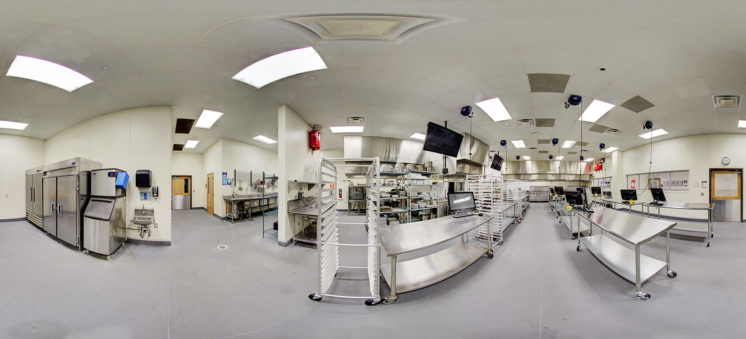 Panoramic view of a commercial kitchen training lab with stainless steel prep tables, industrial ovens and refrigerators, rolling racks, sinks, and multiple computer stations arranged along the counters under bright overhead lighting.
